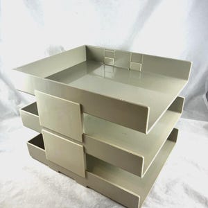 May include: A light gray plastic desk organizer featuring three stacked horizontal trays with open fronts and square-shaped front panels. The unit has a modular design with visible interlocking tabs on the top rear edge for vertical stacking.