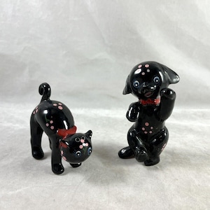 May include: A pair of black ceramic salt and pepper shakers shaped like a cat and a dog. Both are decorated with pink polka dots and red bows.