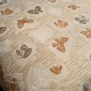 May include: A quilted bedspread with a repeating pattern of butterflies and floral designs in beige, brown, and white. The butterflies have detailed wing patterns, and the fabric has a subtle striped texture.