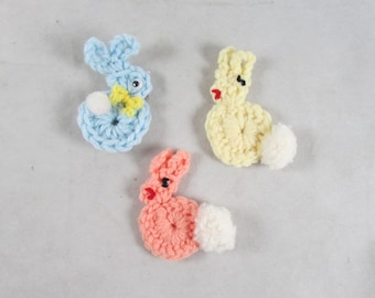 May include: Three crocheted bunny rabbit ornaments. One is light blue, one is yellow, and one is pink. Each bunny has a white pom-pom tail.