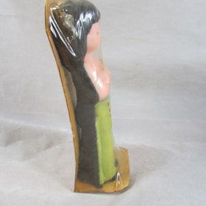 May include: A plastic doll with black hair, a pink torso, and green pants. The doll is packaged in a clear plastic bag with a brown cardboard backing.
