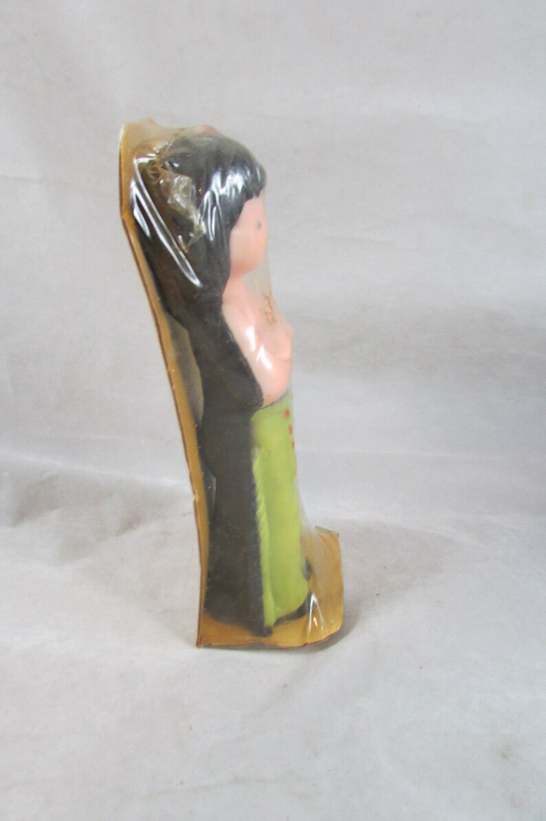 May include: A plastic doll with black hair, a pink torso, and green pants. The doll is packaged in a clear plastic bag with a brown cardboard backing.