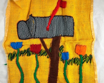 May include: A yellow fabric wall hanging with a gray knitted mailbox, a red flag, and four colorful flowers with green stems. The background is a yellow fabric with a woven texture.