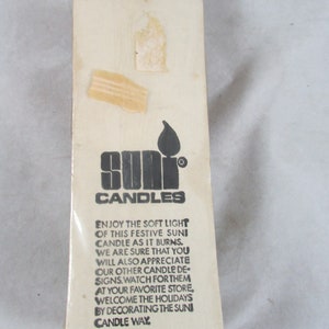 May include: A white cardboard box with black text that reads "Suni Candles". The box features a black and white illustration of a candle flame. The text on the box reads "Enjoy the soft light of this festive Suni candle as it burns. We are sure that you will also appreciate our other candle designs. Watch for them at your favorite store. Welcome the holidays by decorating the Suni candle way. 7 1/2 in. tall candle. Tear or cut along perforated line."