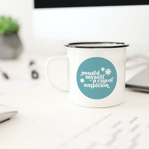May include: White enamel mug with a black rim and a blue circle sticker that says "poured myself a cup of ambition" with white text and a white flower design.