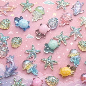 Puede incluir: Una colección de dijes iridiscentes de criaturas marinas, incluyendo estrellas de mar, medusas, tortugas y conchas, sobre un fondo rosa con nubes y estrellas. Los dijes son de varios colores y tamaños.