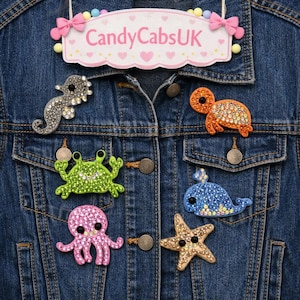 Puede incluir: Una chaqueta de mezclilla azul oscuro con seis parches de criaturas marinas incrustados con cristales: un caballito de mar gris, una tortuga naranja, un cangrejo verde, una ballena azul, un pulpo rosa y una estrella de mar dorada. Letrero CandyCabsUK.