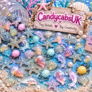 May include: A collection of colorful ocean-themed resin cabochons including starfish, dolphins, jellyfish, and shells. A glass jar spills these charms onto a blue surface. Wooden signs display the text CandycabsUK, Mermaid Vibes Craft Supplies, and Resin Cabochons.