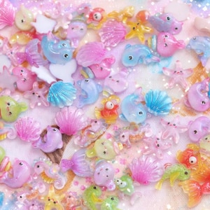 Op de afbeelding: Een verzameling kleurrijke, miniatuurharsbedels met oceaanthema. De collectie bevat dolfijnen, zeesterren, schelpen en vissen in tinten roze, blauw, groen en oranje. Deze kleine, gedetailleerde bedels zijn verspreid over een glinsterende, pastelkleurige achtergrond.