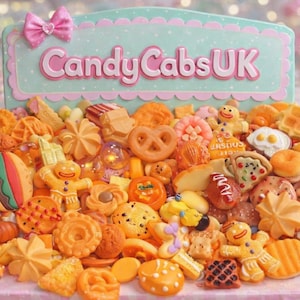 May include: A selection of miniature biscuits and sweets, including gingerbread men and pretzels, arranged on a tray. The sign in the background reads "Candy Cabs UK" with a pink bow. The treats are in various shapes and colours.