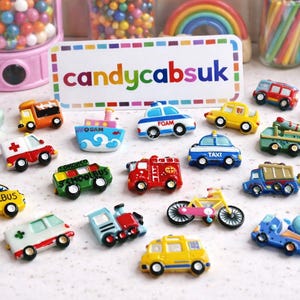 May include: A collection of colorful miniature vehicle-shaped toys, including cars, trucks, a school bus, and a fire truck. The toys are arranged on a white surface, with a sign that reads "candycabsuk" in the background.