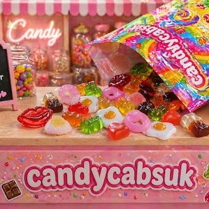 Op de afbeelding: Een kleurrijke zak snoep met het label candycabsuk waaruit diverse gummisnoepjes zoals ringen, lippen en gebakken eieren op een toonbank vallen. Een klein krijtbordje toont de tekst Sweet Treats Just for You! en de merknaam candycabsuk.