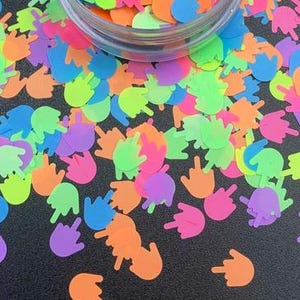 May include: A scattering of neon-coloured glitter in the shape of a hand giving the middle finger. The glitter is on a black surface.