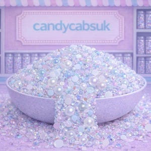 Può includere: Una ciotola traboccante di perline e perle iridescenti, che si riversano su una superficie abbinata. Le perline sono un mix di bianco, azzurro e rosa. Lo sfondo presenta scaffali con barattoli e il testo "candycabsuk".