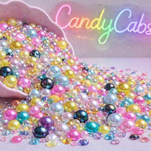 May include: A pink, scalloped bowl overflowing with colorful beads and gems. The beads are in various sizes and colors, including pink, yellow, blue, and iridescent. The text "Candy Cabs UK" is visible in the background.