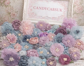 May include: A collection of fabric flowers in shades of dusty blue, mauve, and pale pink, featuring pearl and crystal centers. Behind the flowers sits a white ornate rectangular frame with the text CANDYCABSUK printed in the center.