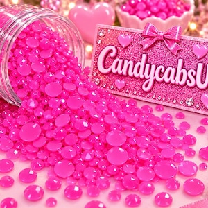 May include: A clear plastic jar spilling out numerous bright pink, round, faceted resin rhinestones. Next to the pile is a rectangular pink glitter sign featuring the text CandycabsUK, decorated with a pink bow, small hearts, and clear crystals.