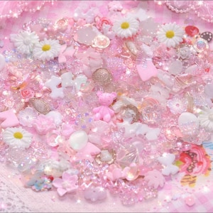 Puede incluir: Una colección de adornos decorativos rosas y blancos, que incluyen corazones, flores y un pequeño oso de peluche. Los elementos están dispersos sobre una superficie rosa y brillante con un borde de encaje, creando una estética caprichosa y encantadora.