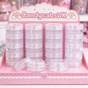 Peut inclure: Contenants en plastique transparents et empilables, présentés sur une étagère rose. Les récipients cylindriques ont des couvercles à visser, parfaits pour ranger de petits objets. L'étagère porte l'inscription "CandycabsUK" avec un arc-en-ciel.