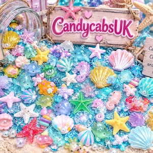Puede incluir: Una colección de cabujones de resina coloridos con conchas, estrellas de mar, caballitos de mar y criaturas marinas en tonos pastel y vibrantes. Un frasco de vidrio derrama estos dijes sobre una superficie de purpurina azul con letreros.