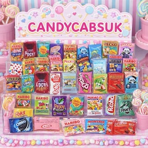Puede incluir: Un expositor rosa con paquetes de caramelos en miniatura, incluyendo Haribo y Pocky. El expositor presenta el texto "CANDYCABSUK" en rosa. Piruletas y otros dulces están dispuestos alrededor, creando una escena colorida.