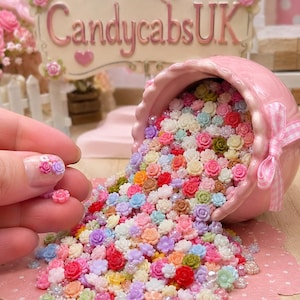 Peut inclure: Un bol en céramique rose renversé sur le côté, déversant une collection de fleurs miniatures en résine de diverses couleurs pastel et vives, dont rose, bleu, jaune et blanc, ainsi que de petites perles transparentes. Une enseigne indique CandycabsUK.
