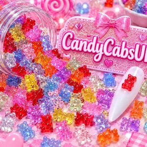 Peut inclure: Une collection de petites breloques translucides et multicolores en forme d'oursons gélifiés, éparpillées autour d'un pot transparent et d'un embout blanc pour nail art. Un panneau rose pailleté avec un nœud rose porte le texte CandyCabsUK.