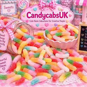 Pode incluir: Um frasco de vidro virado derramando cabochões de resina multicoloridos em forma de minhoca sobre uma superfície. A cena inclui uma placa rosa com o texto CandycabsUK, um pequeno quadro-negro com texto e etiquetas em forma de coração com frases.