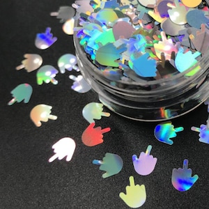 Puede incluir: Un tarro de purpurina holográfica iridiscente con forma de dedos medios. La purpurina está esparcida sobre una superficie negra.