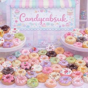 Op de afbeelding: Een verzameling kleurrijke miniatuur donuts met diverse toppings, gepresenteerd op witte taartstandaards en een tafel. De donuts zijn versierd met roze, groene, paarse en chocolade glazuur. Het bord op de achtergrond zegt "Candycabsuk".