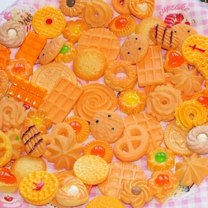 Può includere: Una collezione di biscotti e pasticcini in miniatura arancioni e gialli, tra cui waffle, ciambelle e disegni a spirale. Alcuni hanno condimenti decorativi. Gli articoli sono disposti su una superficie a scacchi rosa e bianca.