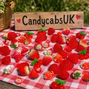 May include: Various red strawberry-shaped sweets with green leaf details, arranged on a red and white checked cloth. A wooden sign with "CandycabsUK" is visible in the background.
