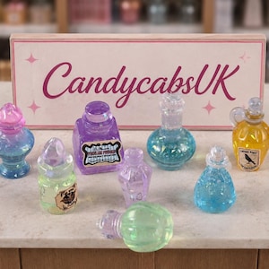 May include: A collection of miniature, colorful potion bottles with glitter inside. The bottles are various shapes and sizes, with pink, blue, green, yellow, and purple hues. The sign behind the bottles reads "CandycabsUK".