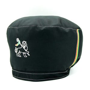 Puede incluir: Boina negra con costuras blancas y una franja colorida. La boina presenta un león bordado y una persona montándolo. La franja incluye colores verde, amarillo y rojo.