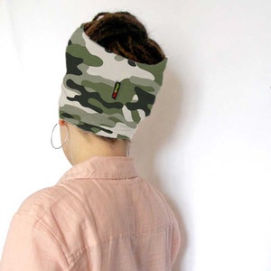 May include: A person wearing a green, white, and black camouflage headband with a red, yellow, and green stripe on the side. The headband is worn over a bun of dreadlocks.