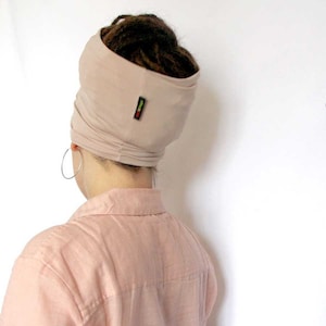 Op de afbeelding: Een beige turban van zachte stof. De turban wordt gedragen door een persoon met dreadlocks. De turban heeft een klein groen, geel en rood label aan de zijkant.
