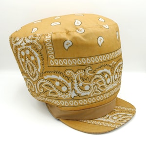 May include: Mustard yellow bandana print cap with a brown brim. The cap features a repeating paisley pattern in white and brown. The cap has a flat top and a short, curved brim. The cap is made of a soft, fabric material.