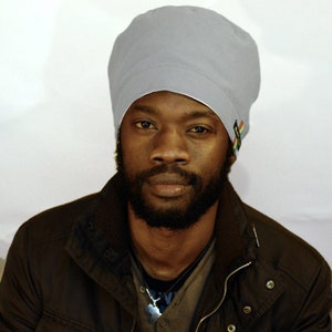 May include: A man with a beard wearing a gray beanie with a colorful design on the side. He is wearing a brown jacket.
