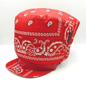 May include: A red bandana-style cap with a white paisley pattern. The cap has a short brim and a rounded crown. The brand name "Hatafaya" is visible on a small tag. The cap is made of a soft fabric.