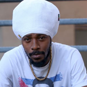 May include: A white waffle-knit tam hat featuring a small black tag with red, yellow, and green stripes on the side. The hat has a rounded, voluminous shape and a textured grid pattern throughout the fabric, providing a distinct look and feel.