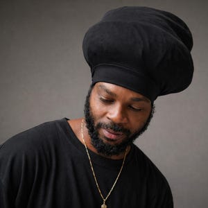May include: A black chef's hat with a rounded crown and a fitted band. The hat is worn by a person wearing a black t-shirt and a gold chain necklace. The background is a solid gray.