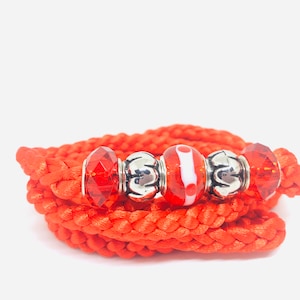May include: A wrapped orange bracelet with decorative beads. The bracelet is made of a thick, braided cord. The beads include faceted red crystals, silver-colored metal beads, and a red and white patterned glass bead.