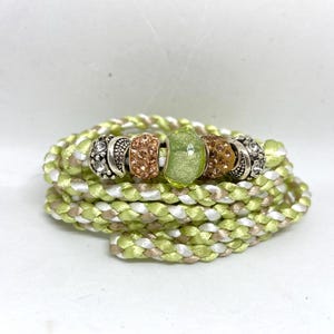May include: A braided bracelet in shades of green, white, and tan, adorned with decorative beads. The bracelet features a central green bead and several silver and bronze-colored accent beads. The bracelet is coiled on a white surface.