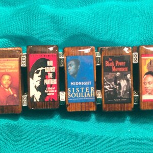 May include: A wooden bracelet with six charms featuring images of books and magazines. The charms include covers of "The Autobiography of Malcolm X", "War Against the Panthers", "Midnight Sister Souljah", "The Black Power Movement", and "Time".