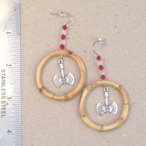 May include: A pair of handmade earrings featuring bamboo hoops with silver axe charms. Red and clear beads dangle from the silver earring hooks. The earrings are displayed on a neutral surface.