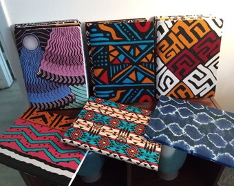 May include: Six different notebooks with colorful patterned covers. The covers feature geometric designs, abstract patterns, and tie-dye effects. The notebooks are stacked on a wooden surface.
