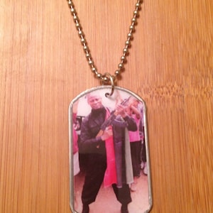 May include: A silver chain necklace with a silver dog tag pendant. The dog tag has a photo of a man holding a rifle. The man is wearing a black jacket and black pants. The photo is in color.