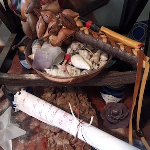 May include: A collection of spiritual items including a gourd with a geometric design, a bowl of seashells, a rolled paper with text, and a leather-wrapped instrument. The items are arranged on a shelf with a dark wooden frame.