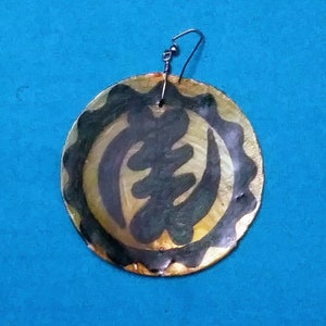 May include: A gold and black circular earring with a black Adinkra symbol, the Sankofa, painted on it. The earring is made of a natural material, possibly shell.
