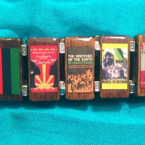 May include: A wooden bracelet with six charms, each featuring a different image. The charms are decorated with red, green, black, yellow, and white colours. The images include a flag, a book cover, a group of people, a woman's face, and a book cover with the word "YURUGU".